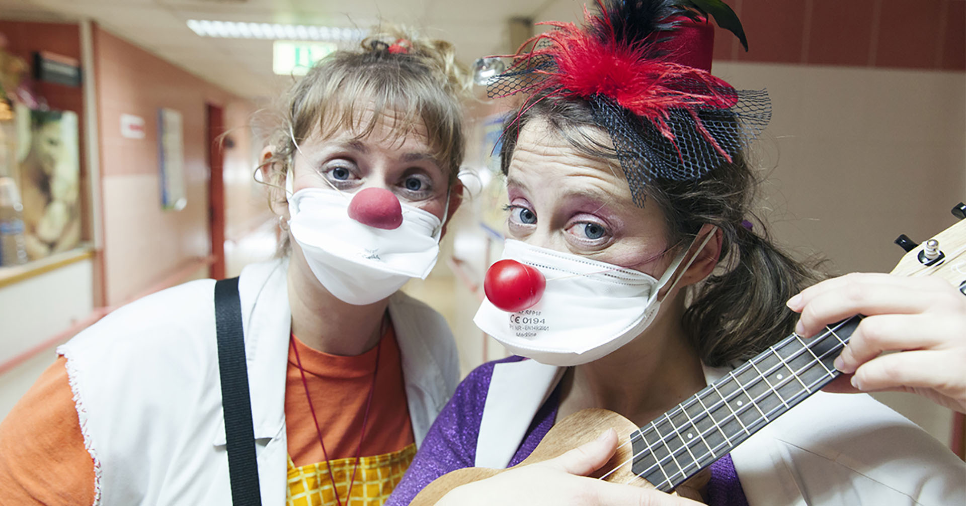 Dos payasas de hospital con mascarillas y narices de payaso mirando a cámara y tocando el ukelele